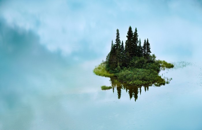 Island with trees reflected in calm water under cloudy sky