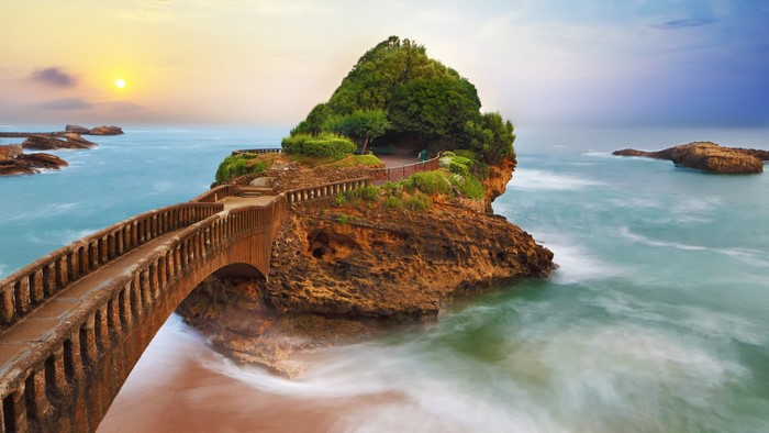 Bridge to a rocky island with trees and the sun over the sea