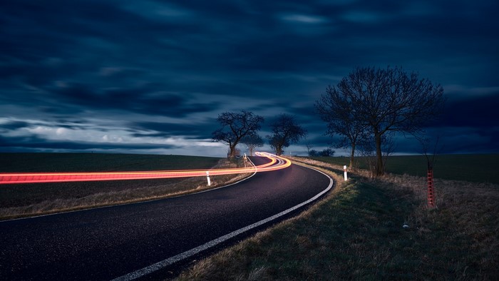Road with light trails at night under a cloudy sky