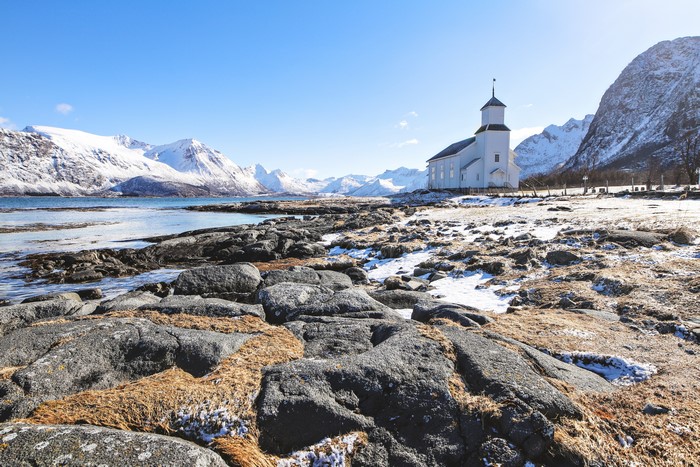 White church on a rocky beach with snow-capped mountains in Norway