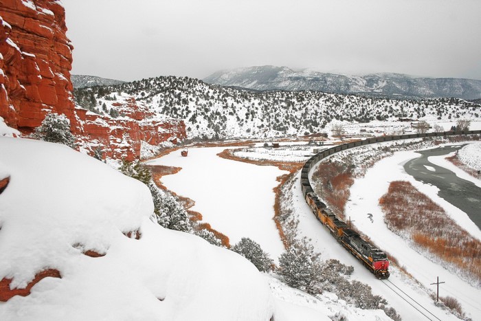 Train traveling through a snowy river valley with red rocks