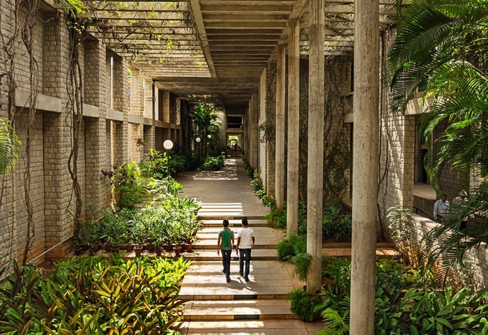 Two men walk down a stone pathway lined with plants and columns.