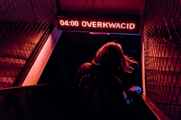 Person walking down stairs under "04:00 OVERKWACID" sign