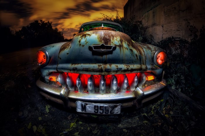 Rusted vintage car with glowing red headlights and grille at night