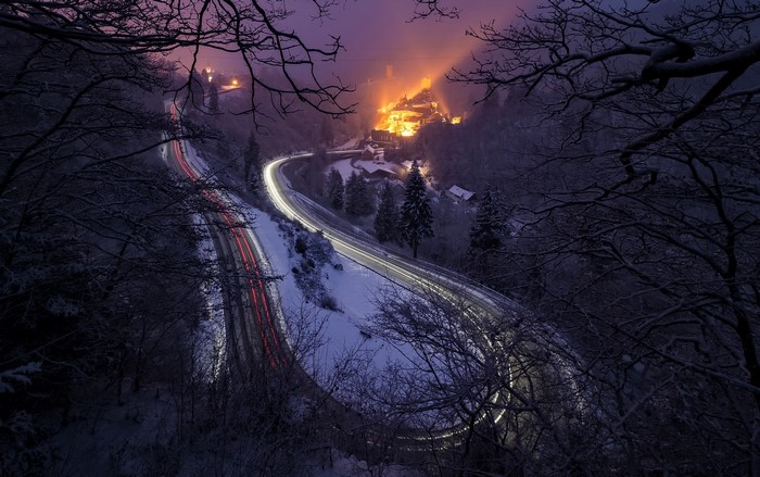 Long exposure of a snowy road with car lights and a village at night