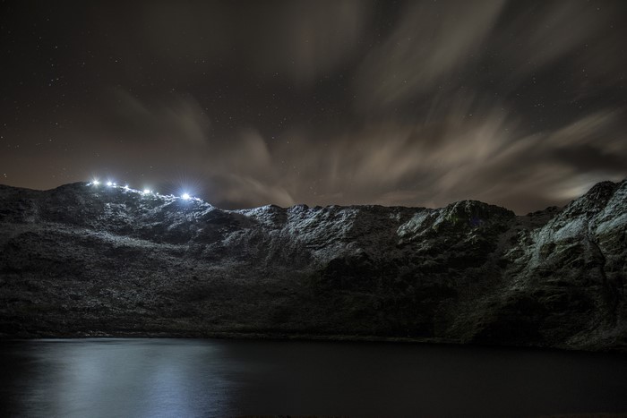 Night landscape with stars, clouds, and lights on mountains over water