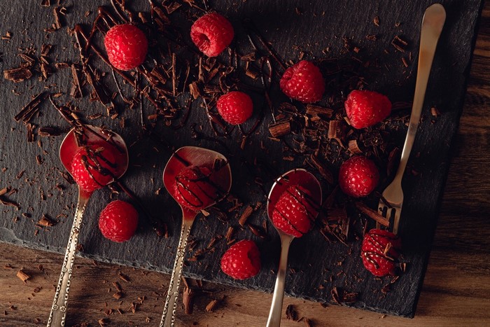 Raspberries and chocolate shavings on a slate board with spoons and a fork