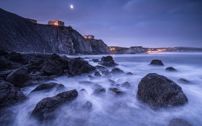 Long exposure of rocky coastline with buildings and moon