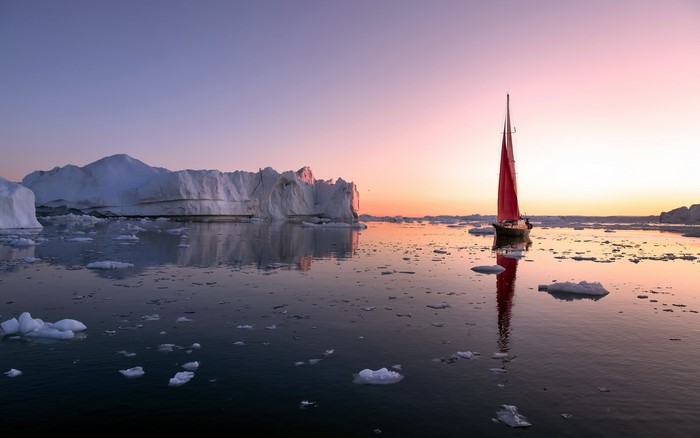 Sailboat with red sail among icebergs at sunset