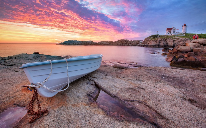 White rowboat on rocky shore with lighthouse at sunset