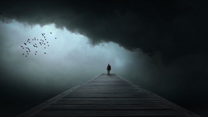 A lone figure walks on a pier under a dark sky with birds