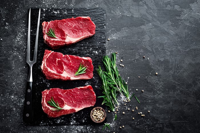 Raw steaks with rosemary and spices on a dark surface