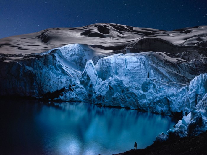 Man silhouetted by a glowing glacier lake under starry night sky