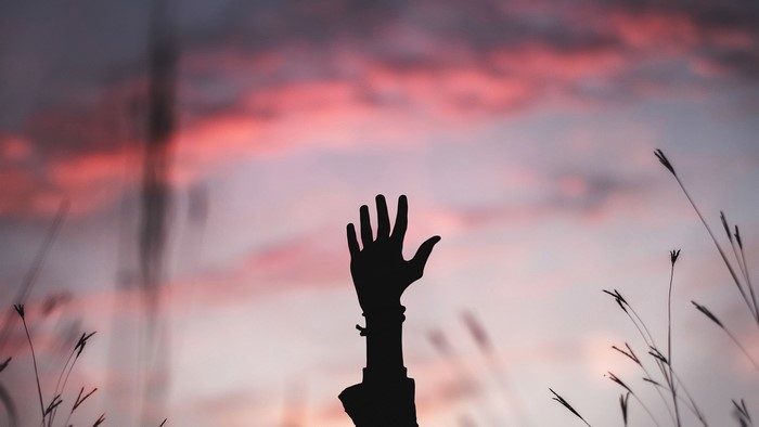 Silhouette of a raised hand against pink clouds at dusk