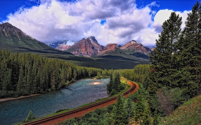 Train tracks curve alongside a river with mountains and forest