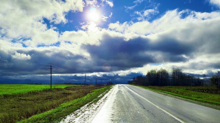 Road through a green field under a cloudy sky with sun