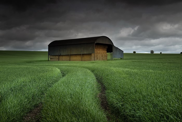Barn with hay bales in a green field under dark clouds
