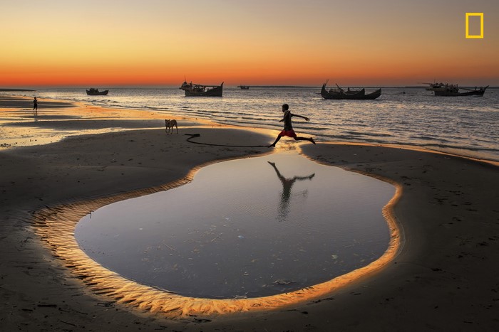 Boy jumping over puddle on beach at sunset with boats