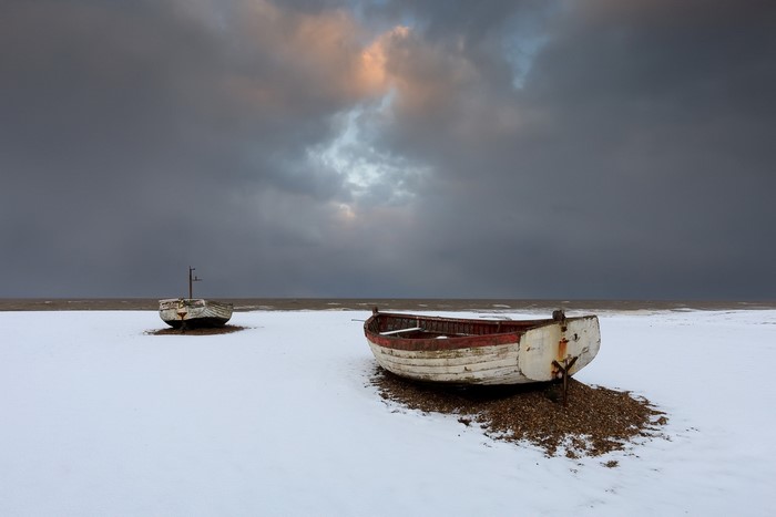 Two old boats on a snowy beach under a cloudy sky