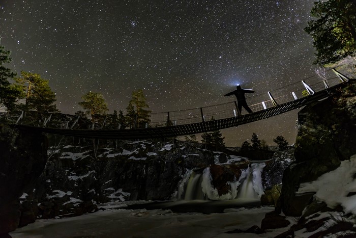 Man on suspension bridge under starry sky with waterfall