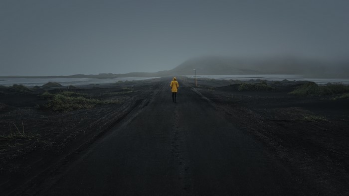 Person in yellow raincoat walking on black sand road