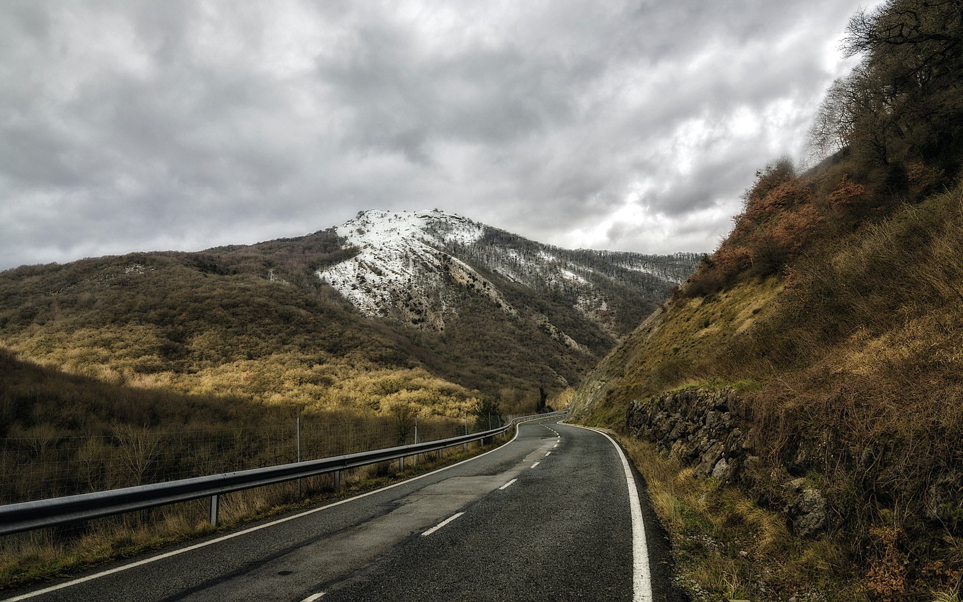 Winding mountain road with snow-capped peak under cloudy sky HD Wallpaper
