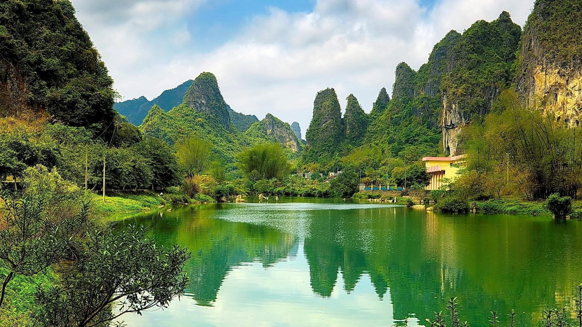Green lake reflecting karst mountains and trees under a cloudy sky HD Wallpaper