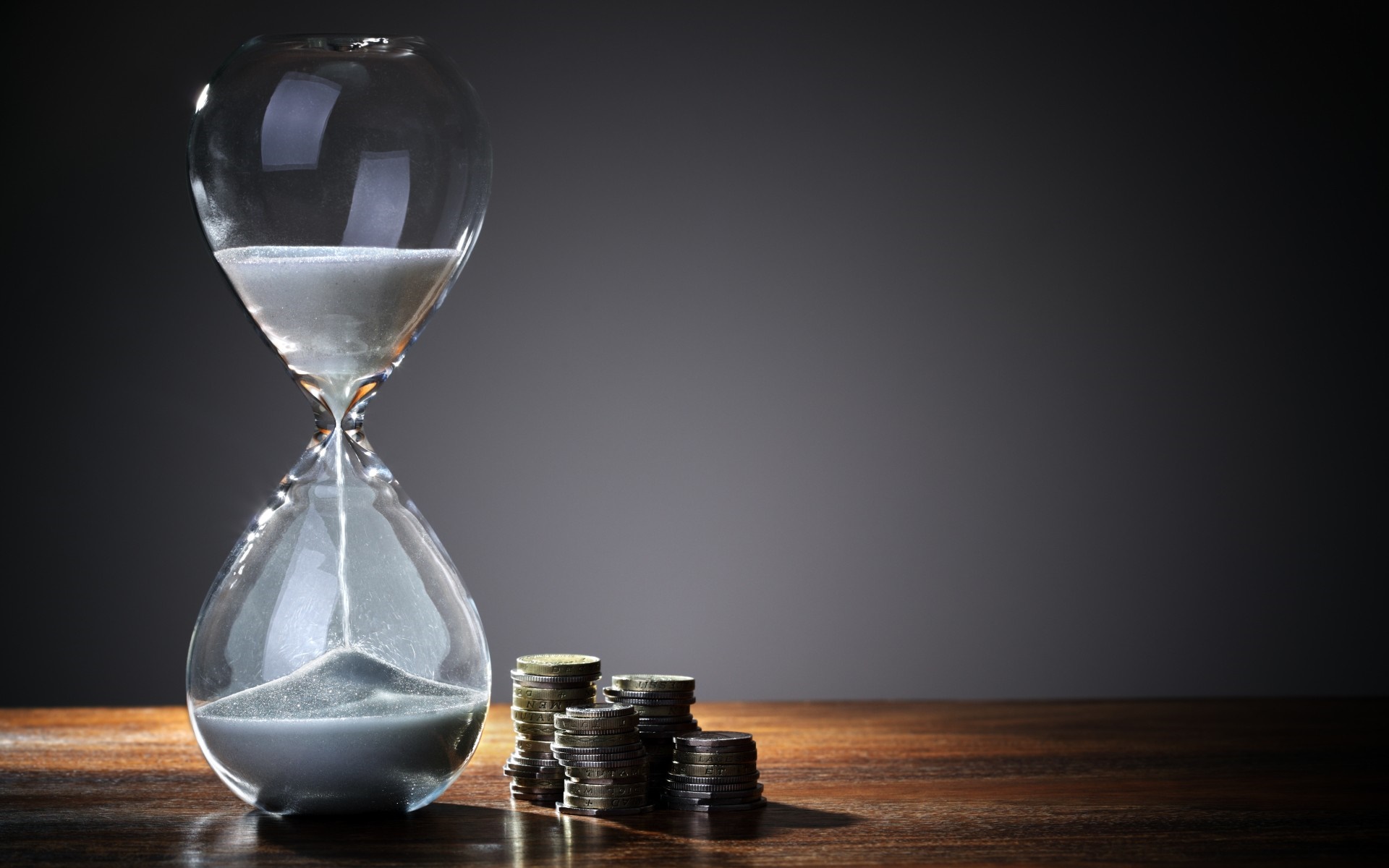 Hourglass with sand and stacks of coins on a wooden table HD Wallpaper