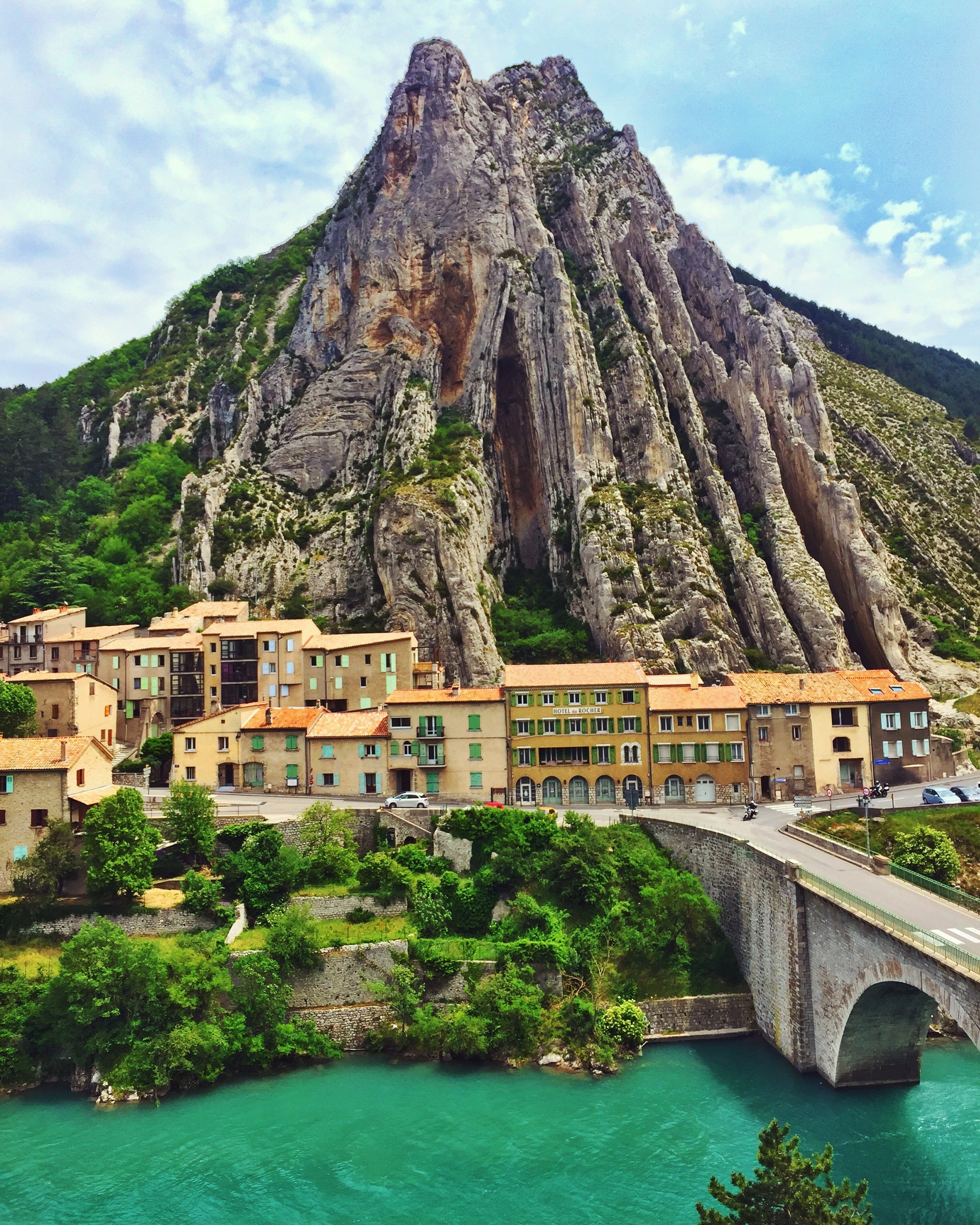 Village nestled below a dramatic mountain by a turquoise river in France HD Wallpaper