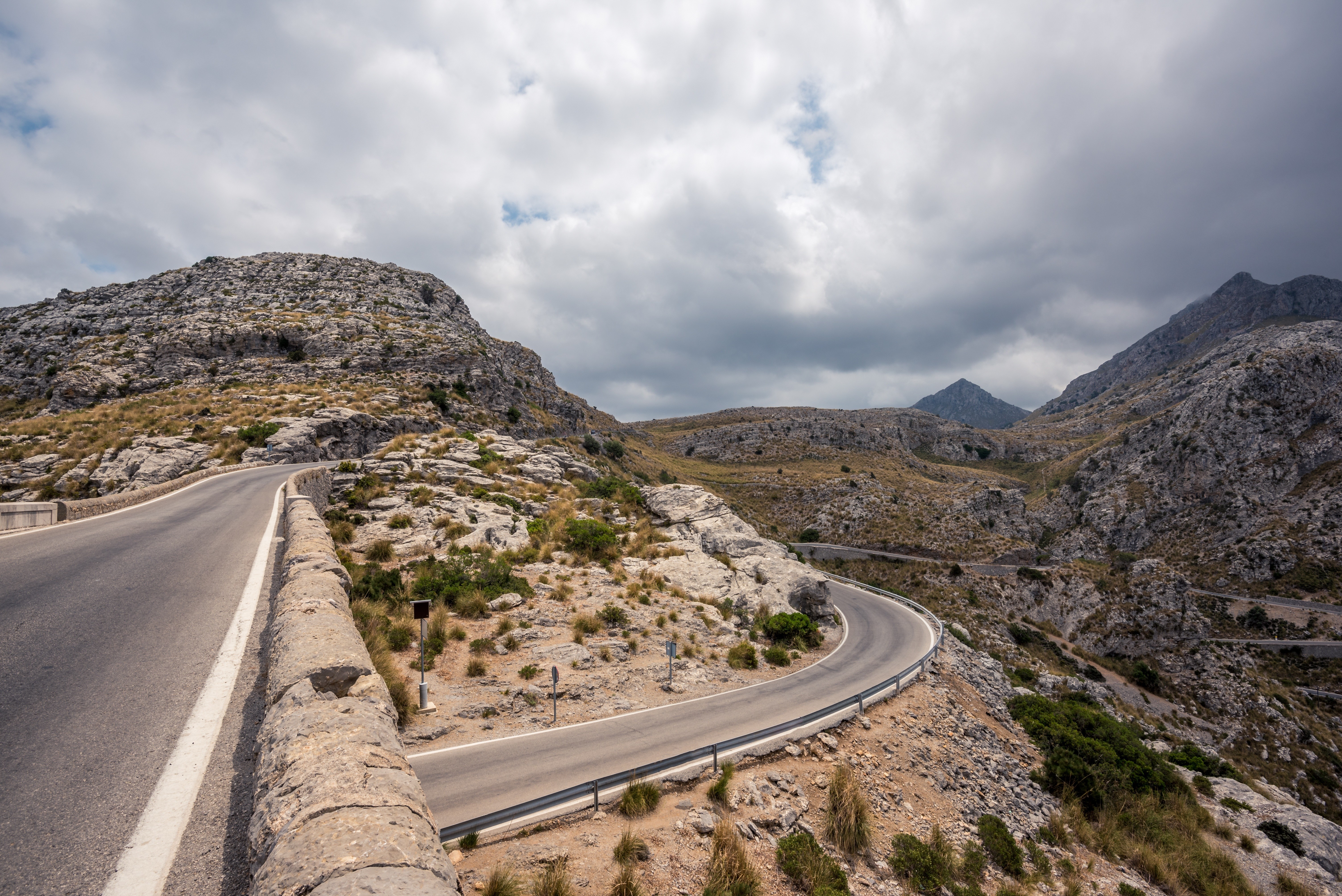 Winding mountain road through rocky terrain under cloudy sky HD Wallpaper