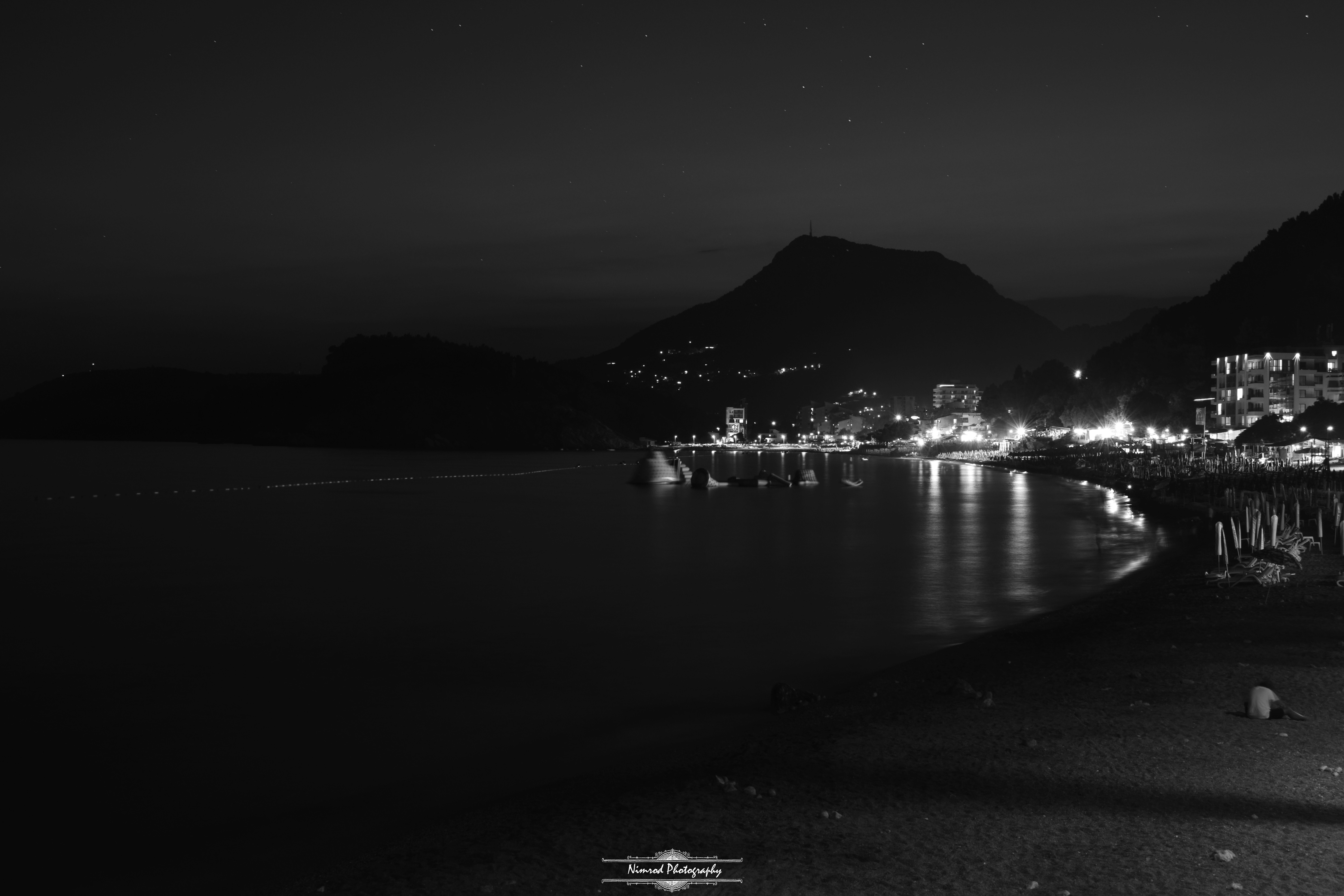 Night view of a coastal town with illuminated buildings and a calm sea HD Wallpaper