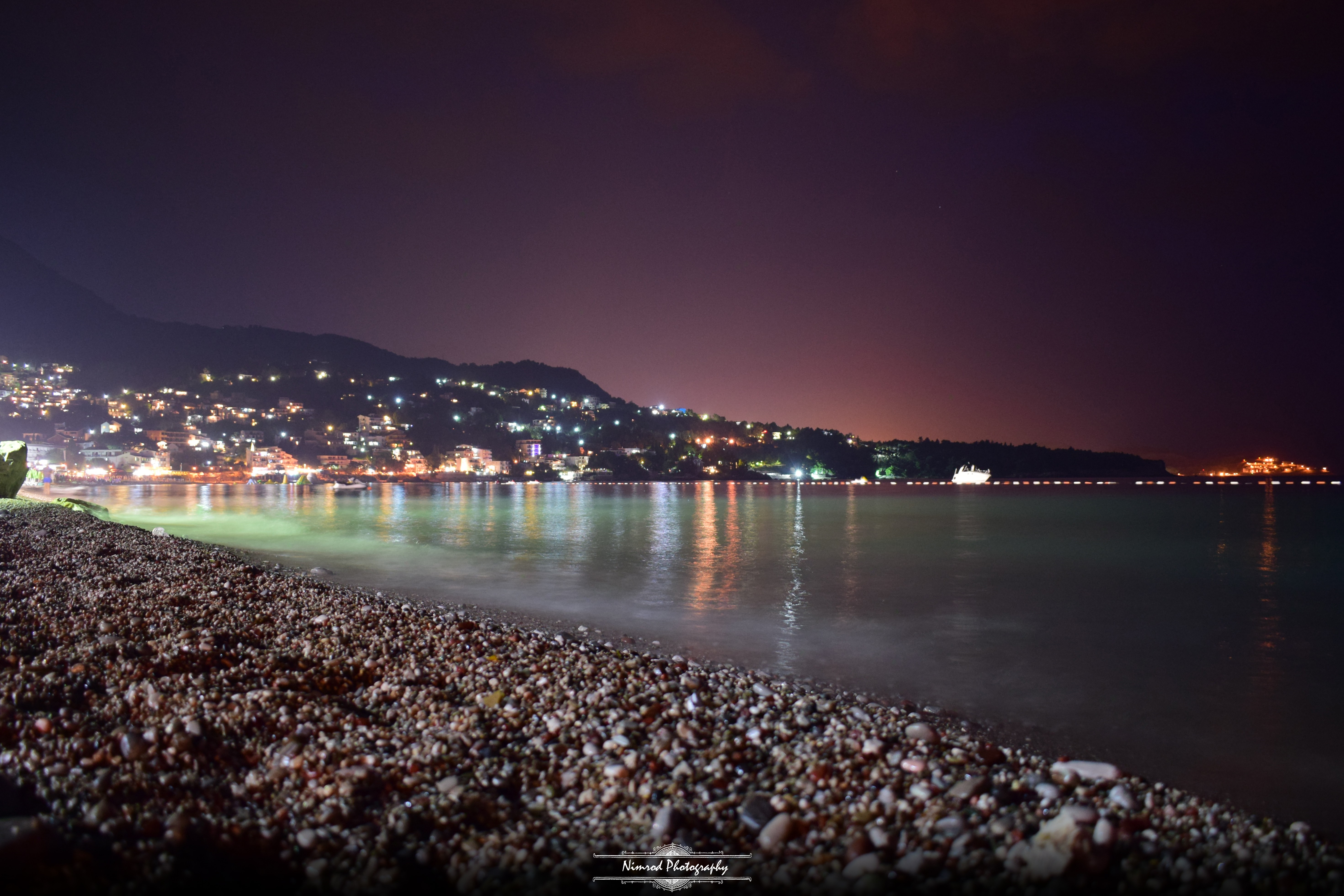 City lights reflecting on the sea at night from a pebble beach HD Wallpaper