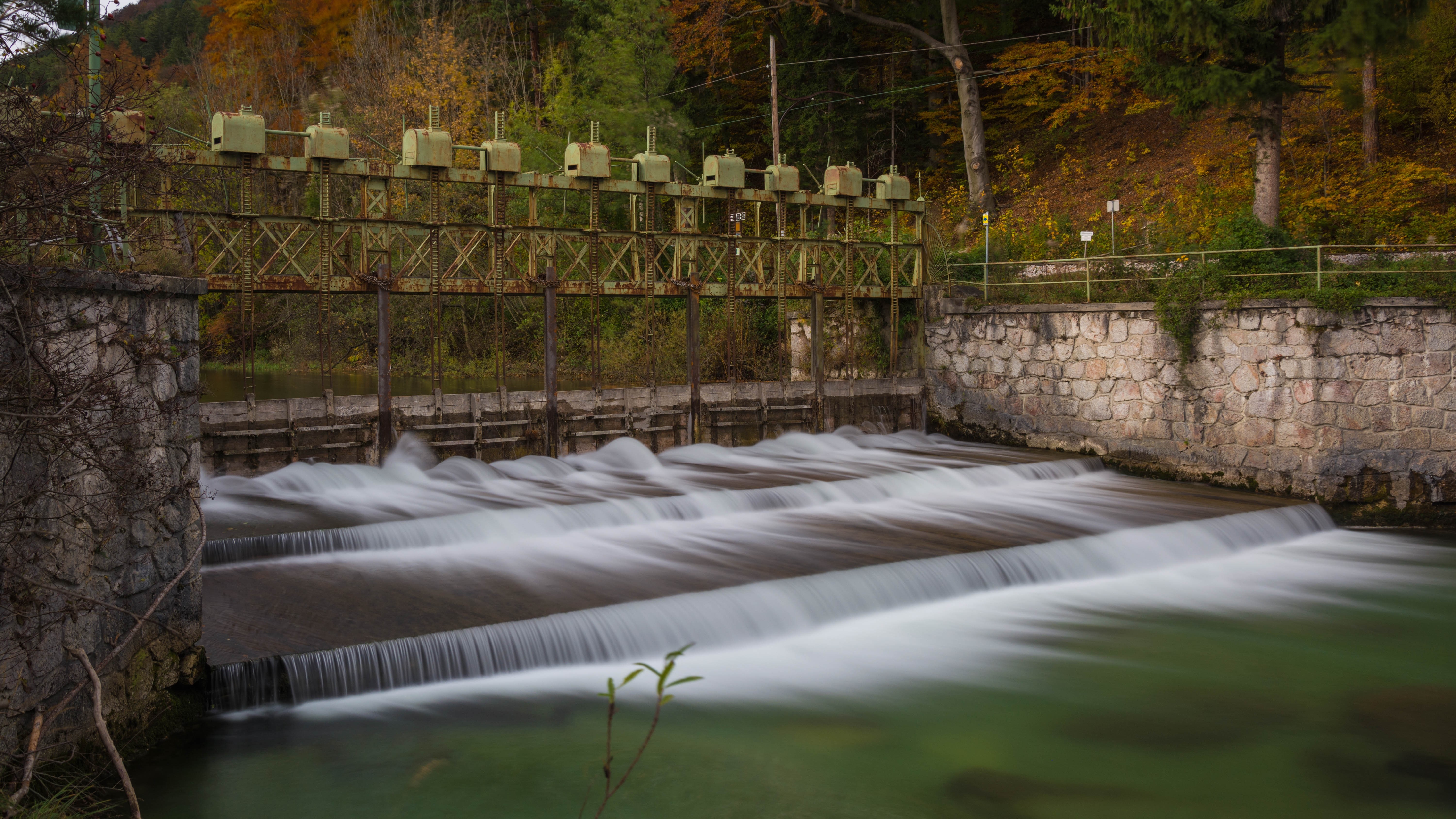 Water flowing over a dam with autumn trees in the background HD Wallpaper