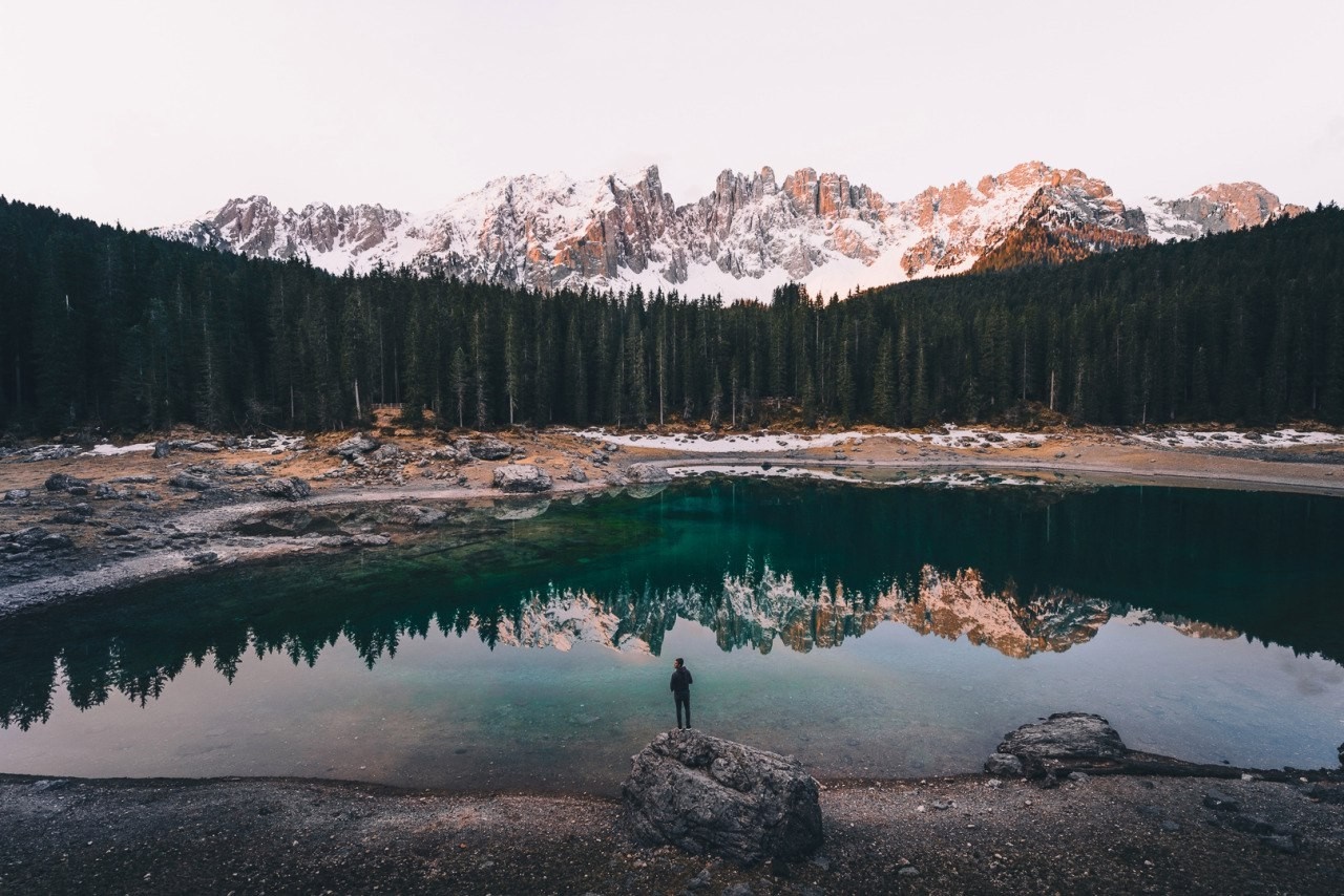Man standing by a lake with snow-capped mountains and forest reflection HD Wallpaper