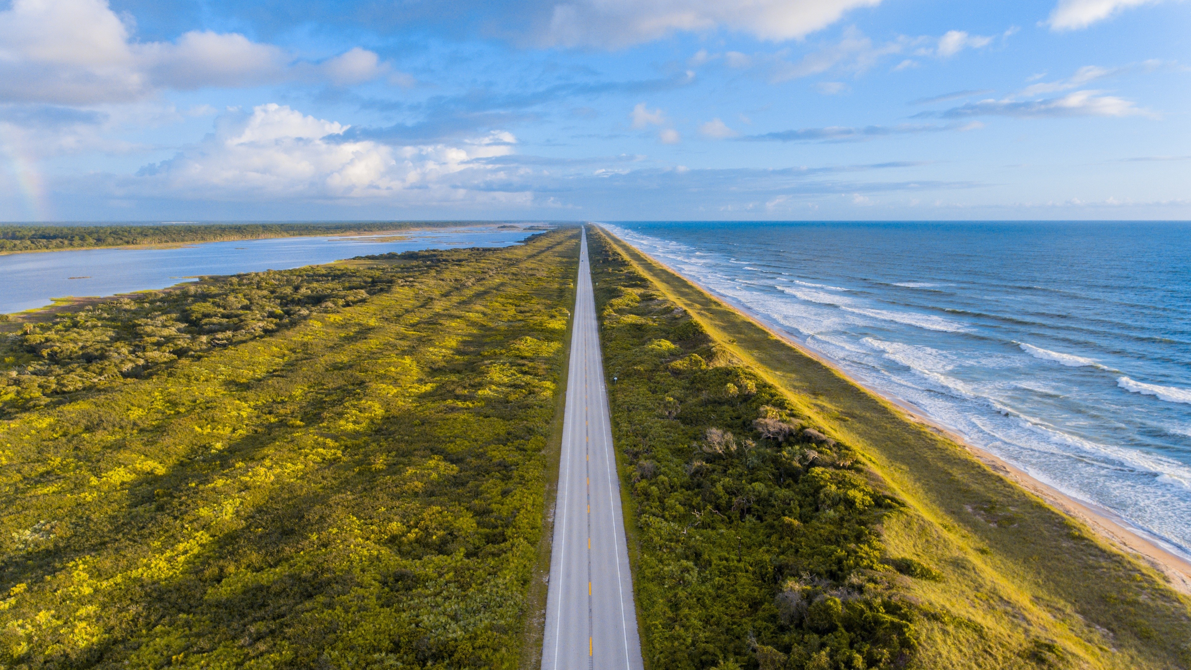 Straight road between ocean and lake with vegetation HD Wallpaper