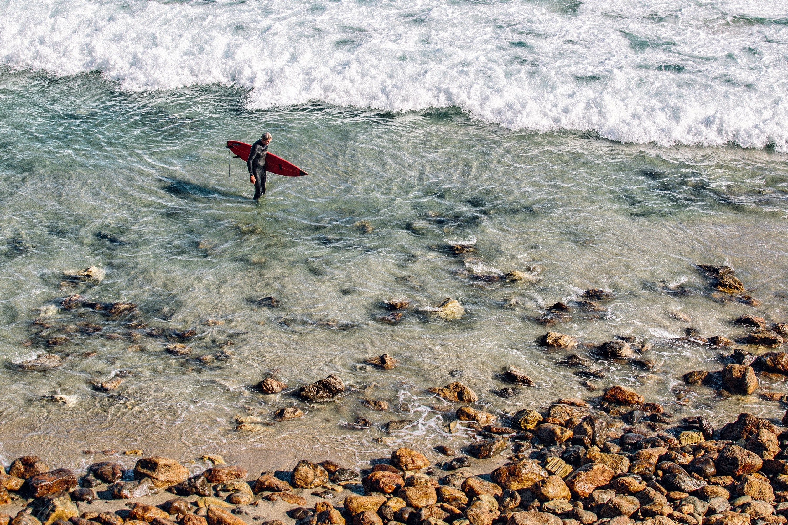 Surfer in wetsuit holding surfboard in shallow water with waves HD Wallpaper