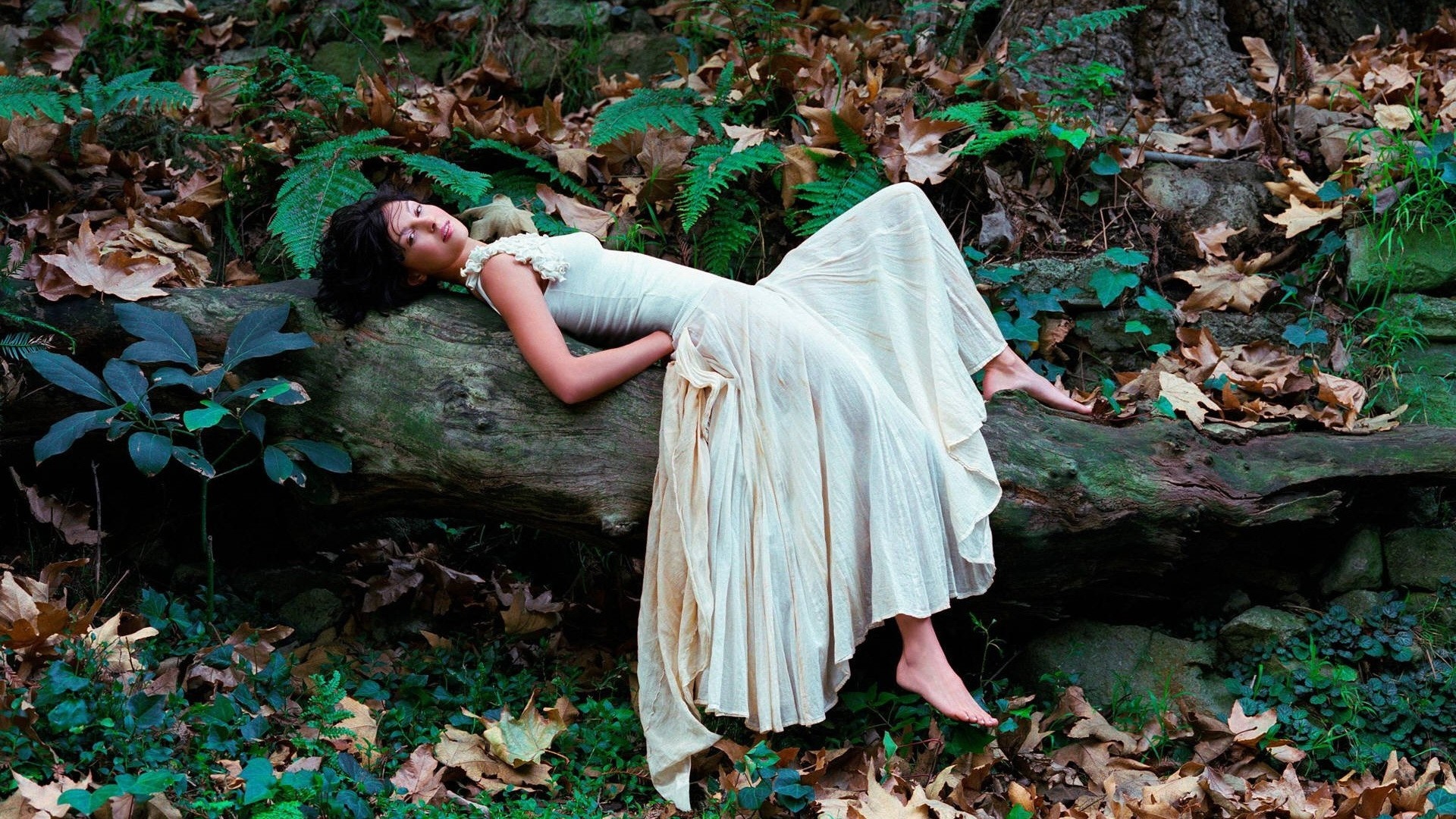 Woman in a white dress lying on a fallen tree in a forest HD Wallpaper