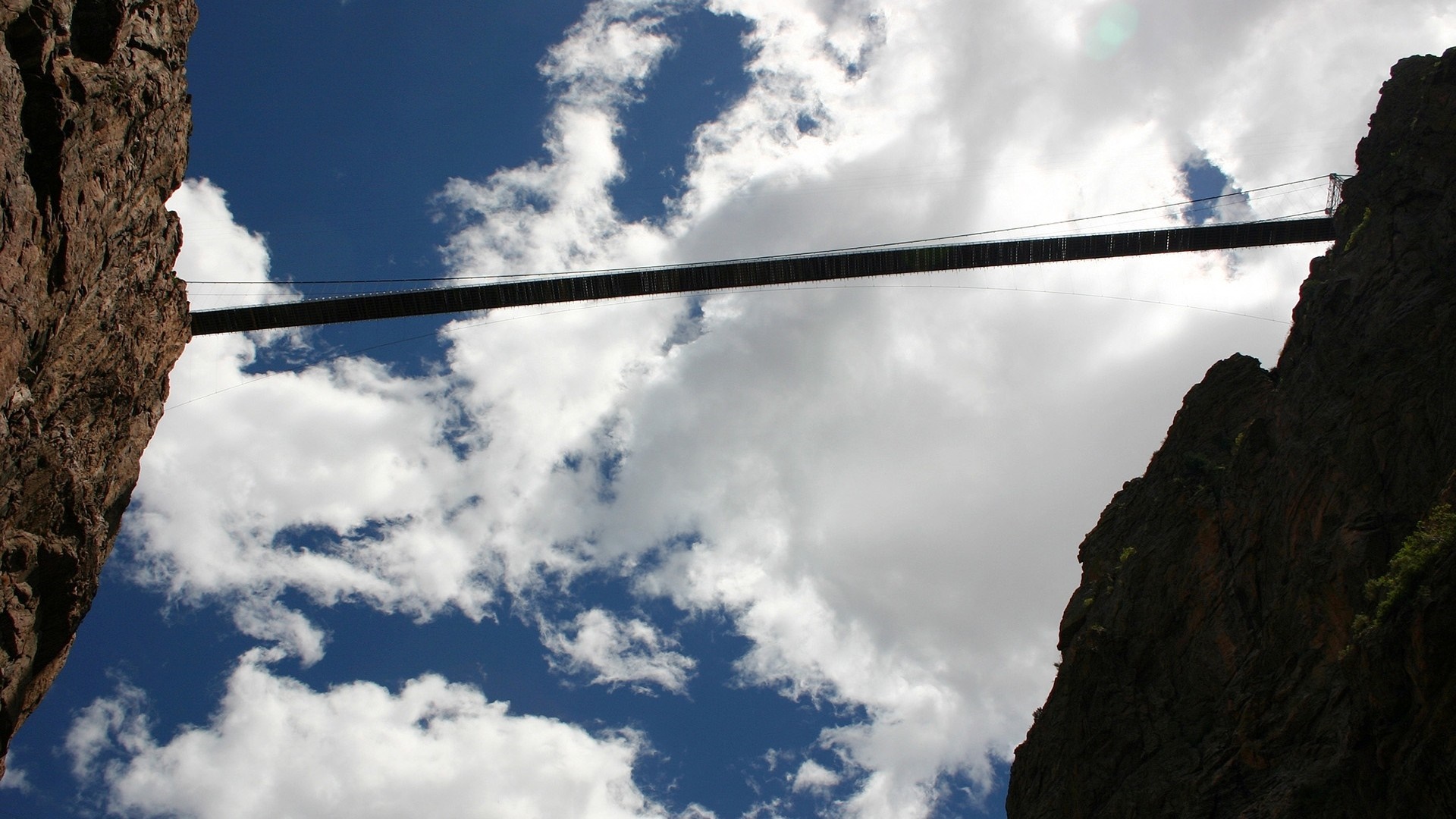 Suspension bridge between rocky cliffs under a cloudy sky HD Wallpaper
