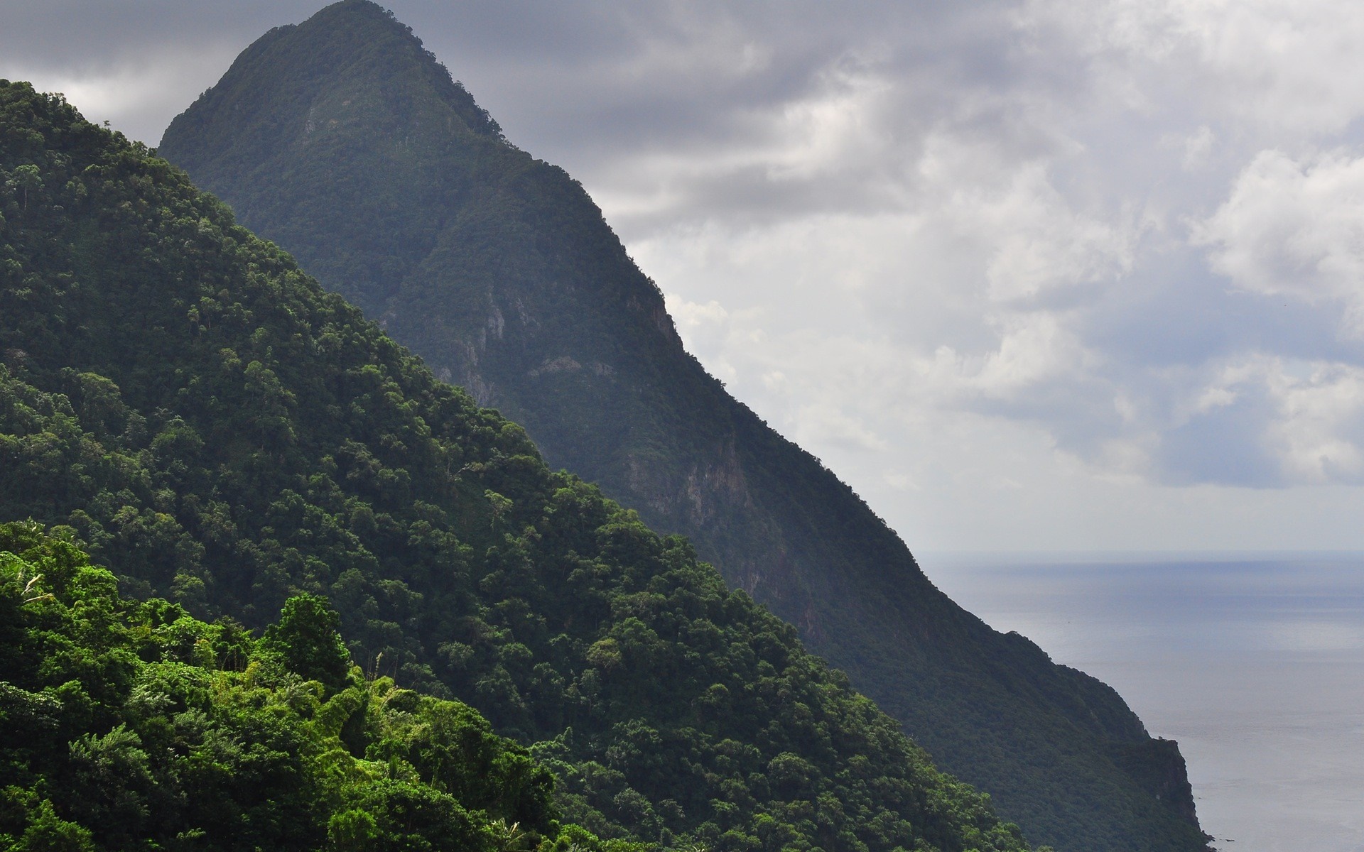 Two lush green mountains meet the sea under a cloudy sky HD Wallpaper