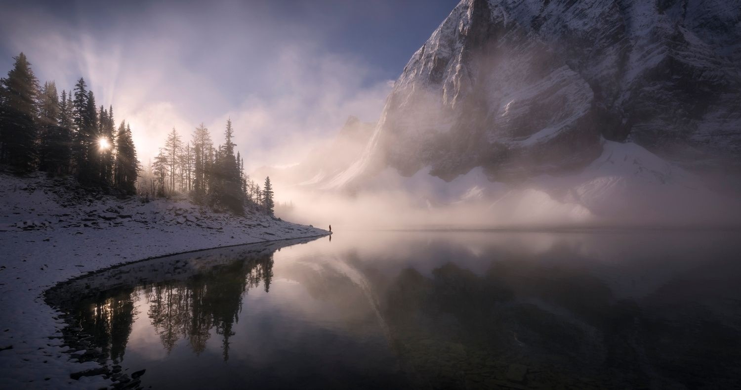 Man standing by a misty lake with snow-covered trees and mountains HD Wallpaper