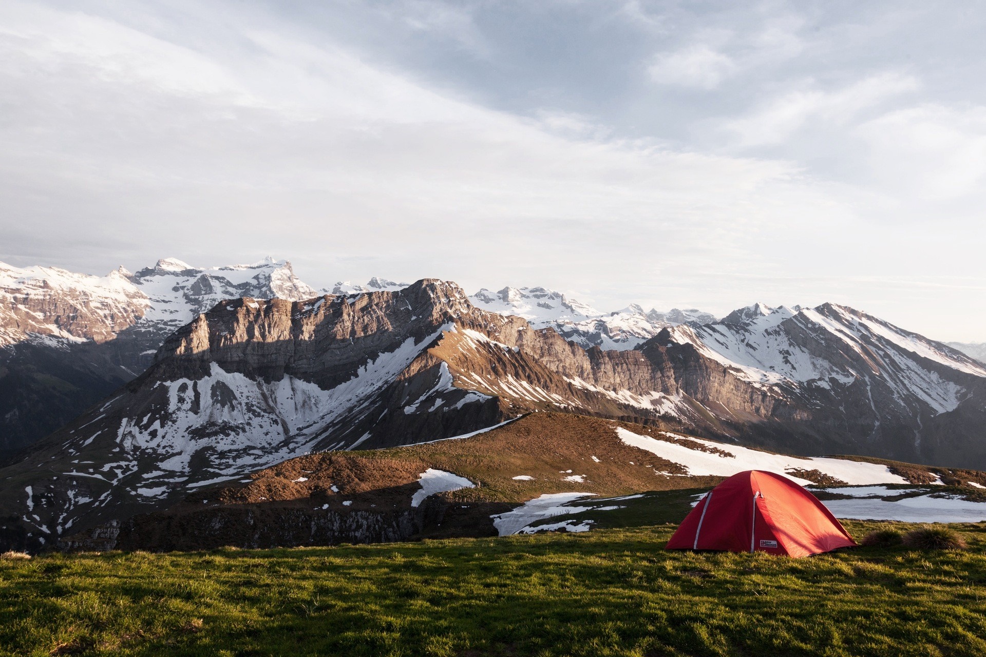 Red tent pitched on grassy mountain slope with snow-capped Alps in background HD Wallpaper