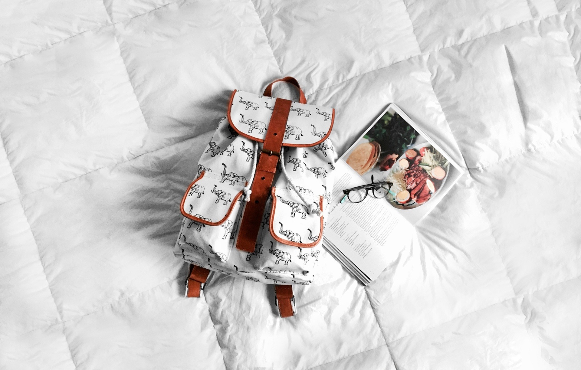 Backpack with elephant pattern on a white comforter with a magazine and glasses HD Wallpaper
