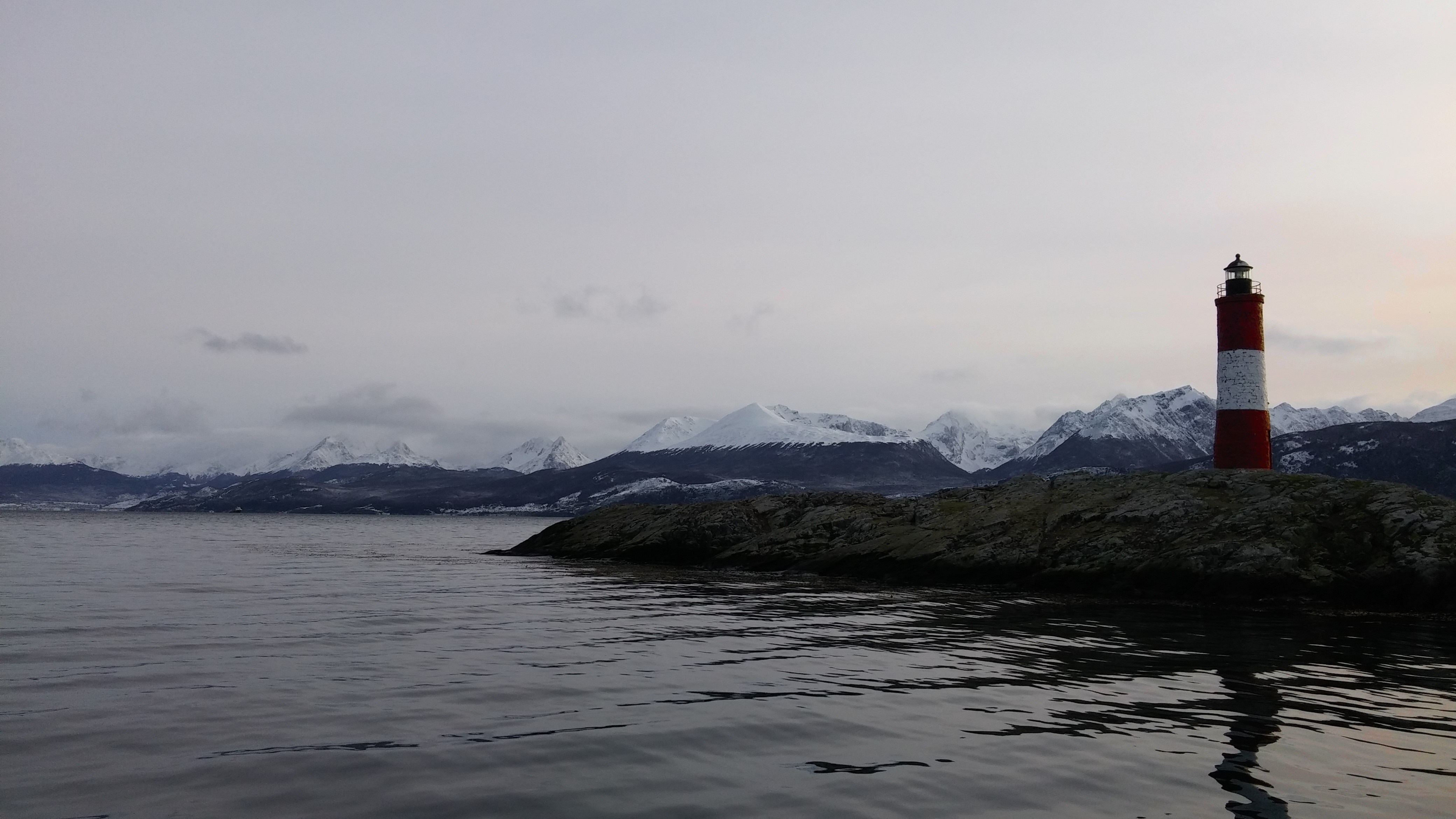 Red and white lighthouse on rocky shore with snow-capped mountains HD Wallpaper