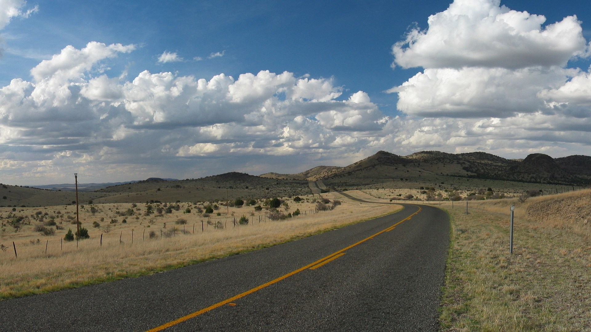 Road through a dry, hilly landscape under a cloudy sky HD Wallpaper