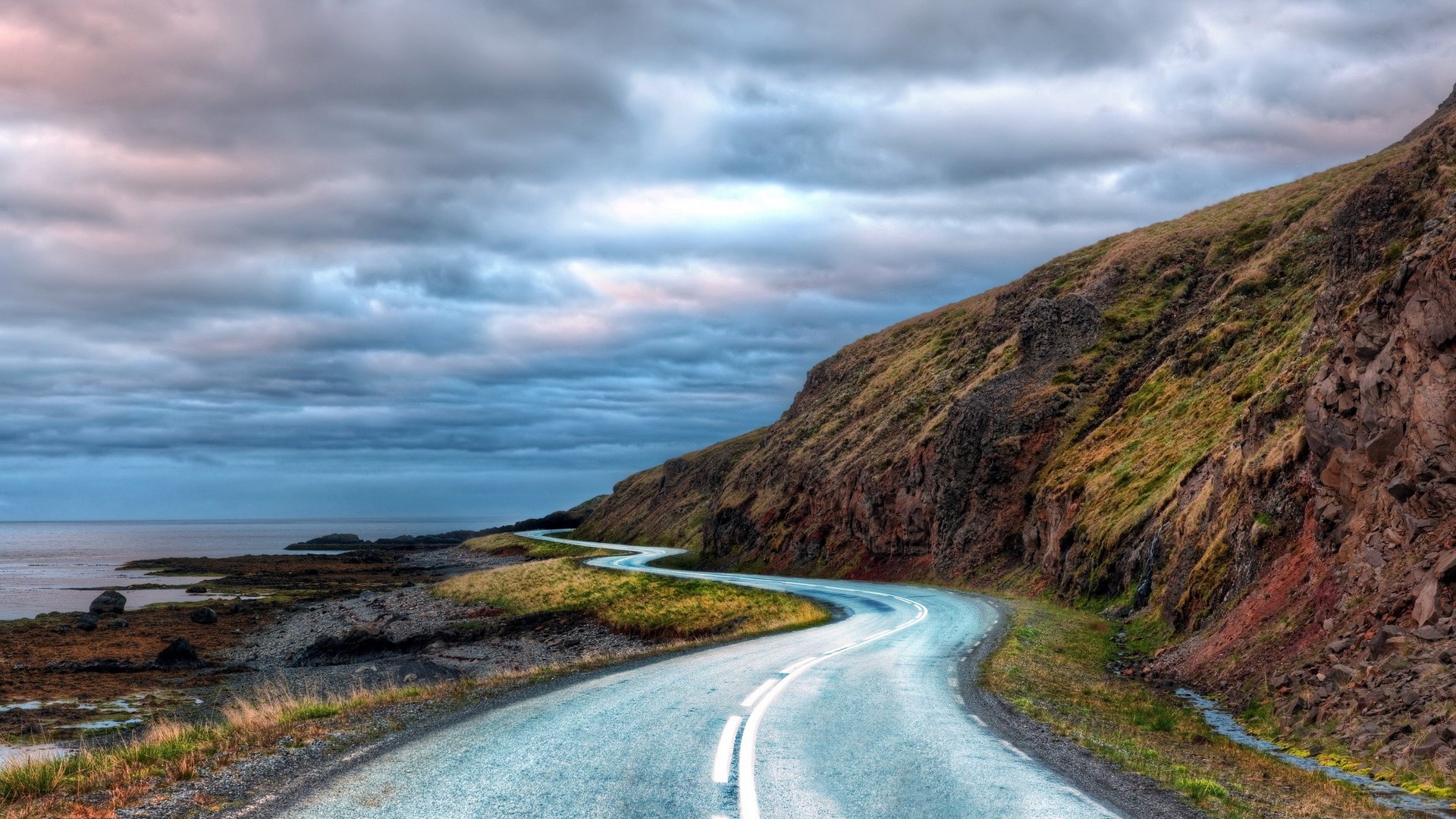 Winding road along a rocky coastline under cloudy sky HD Wallpaper