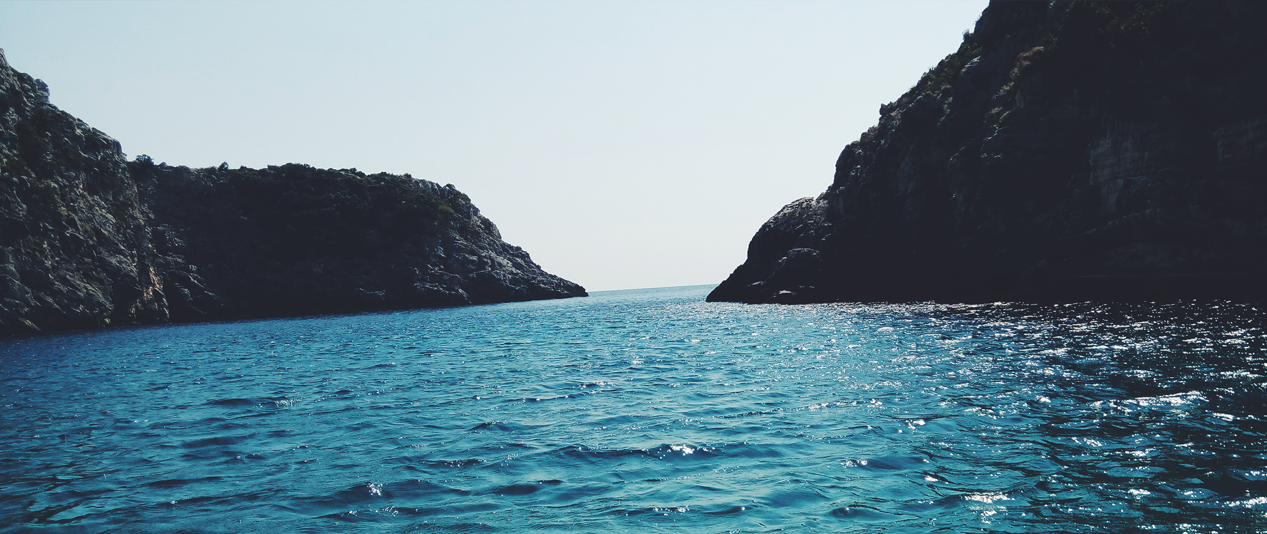 Ocean between two rocky cliffs under a clear sky HD Wallpaper