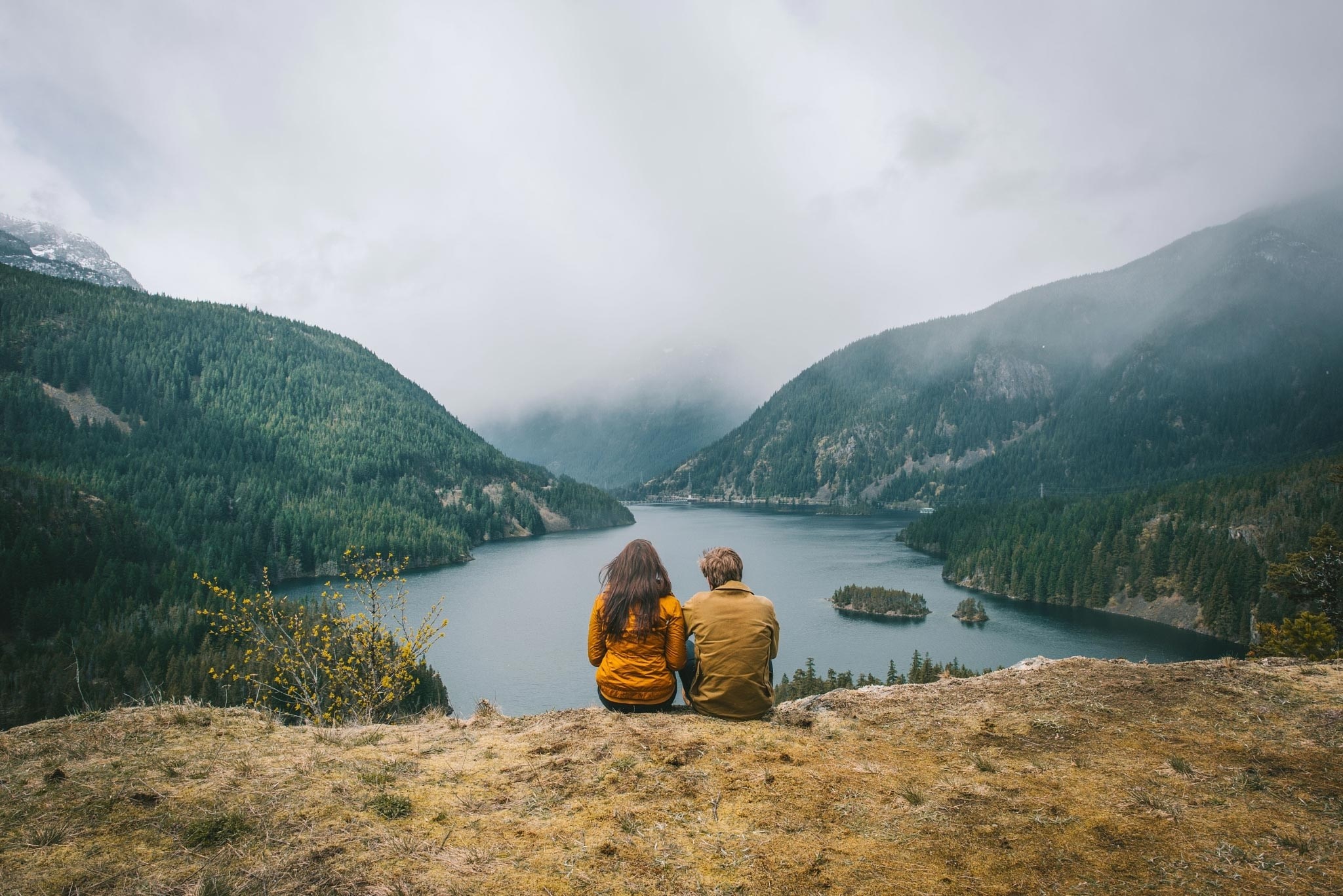 Couple sitting on a cliff overlooking a misty mountain lake HD Wallpaper