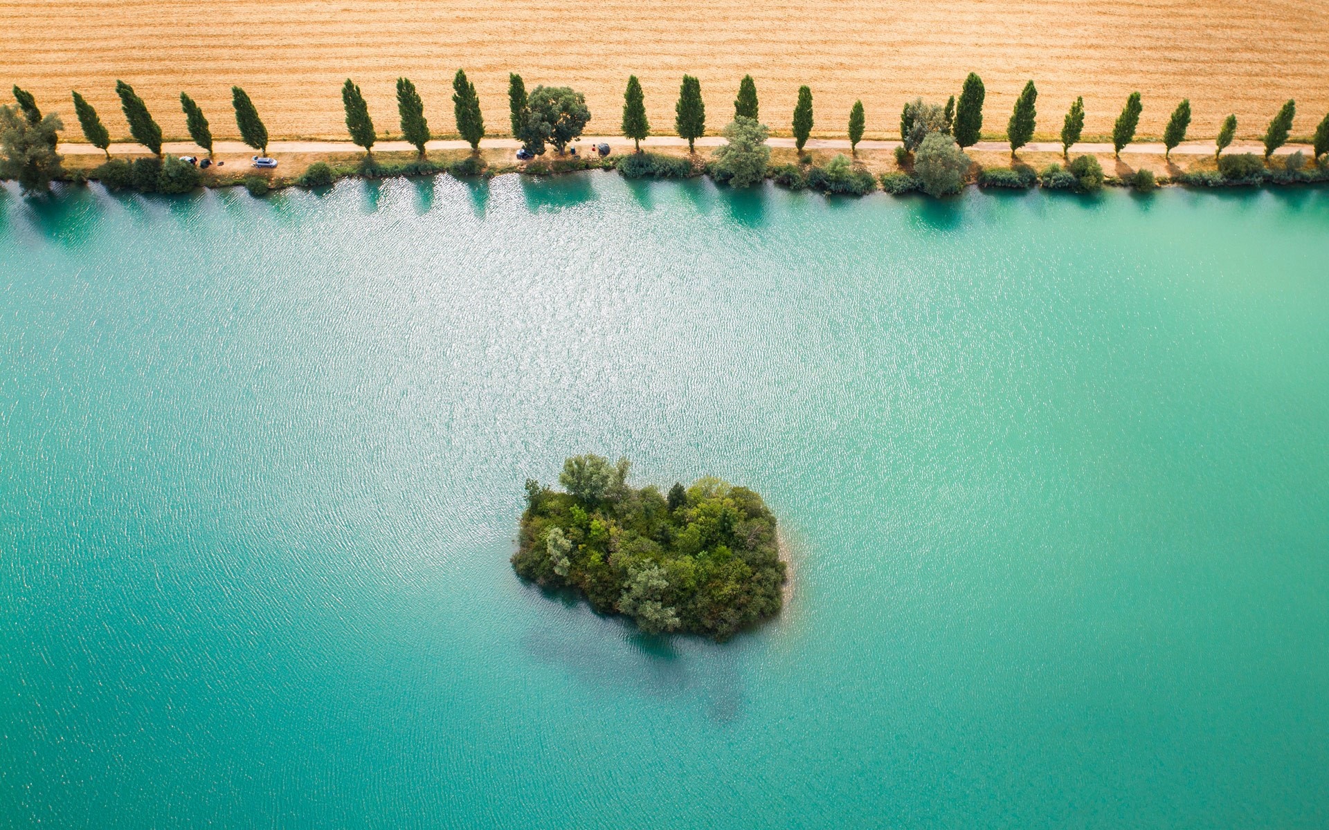 Aerial view of a small green island in a turquoise lake with a field and trees HD Wallpaper