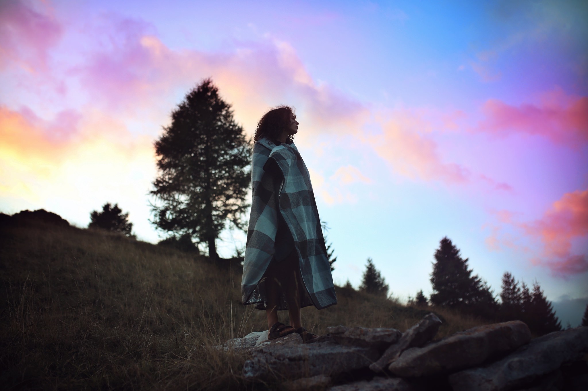 Woman wrapped in blanket on rocky hill at sunset HD Wallpaper