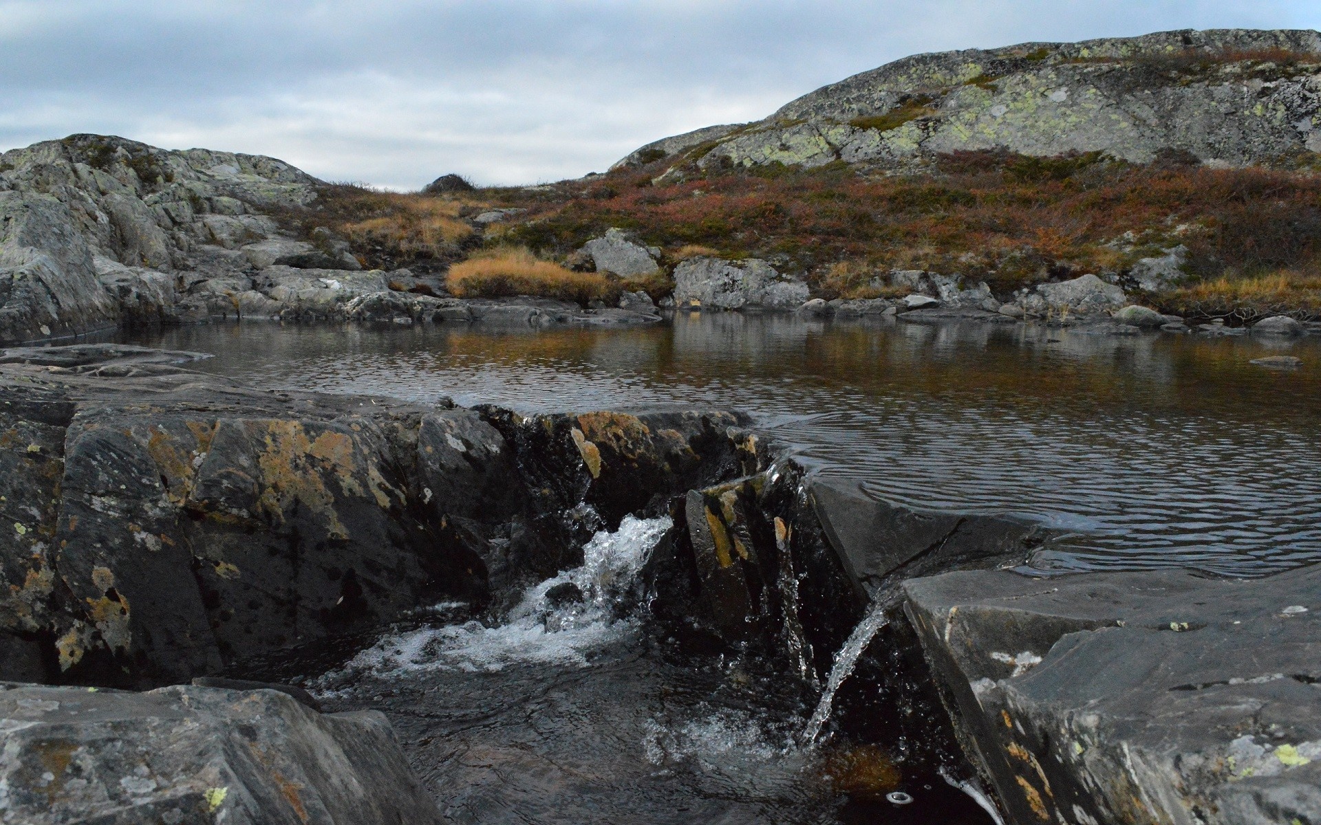 Rocky stream flowing into a calm lake with hills HD Wallpaper